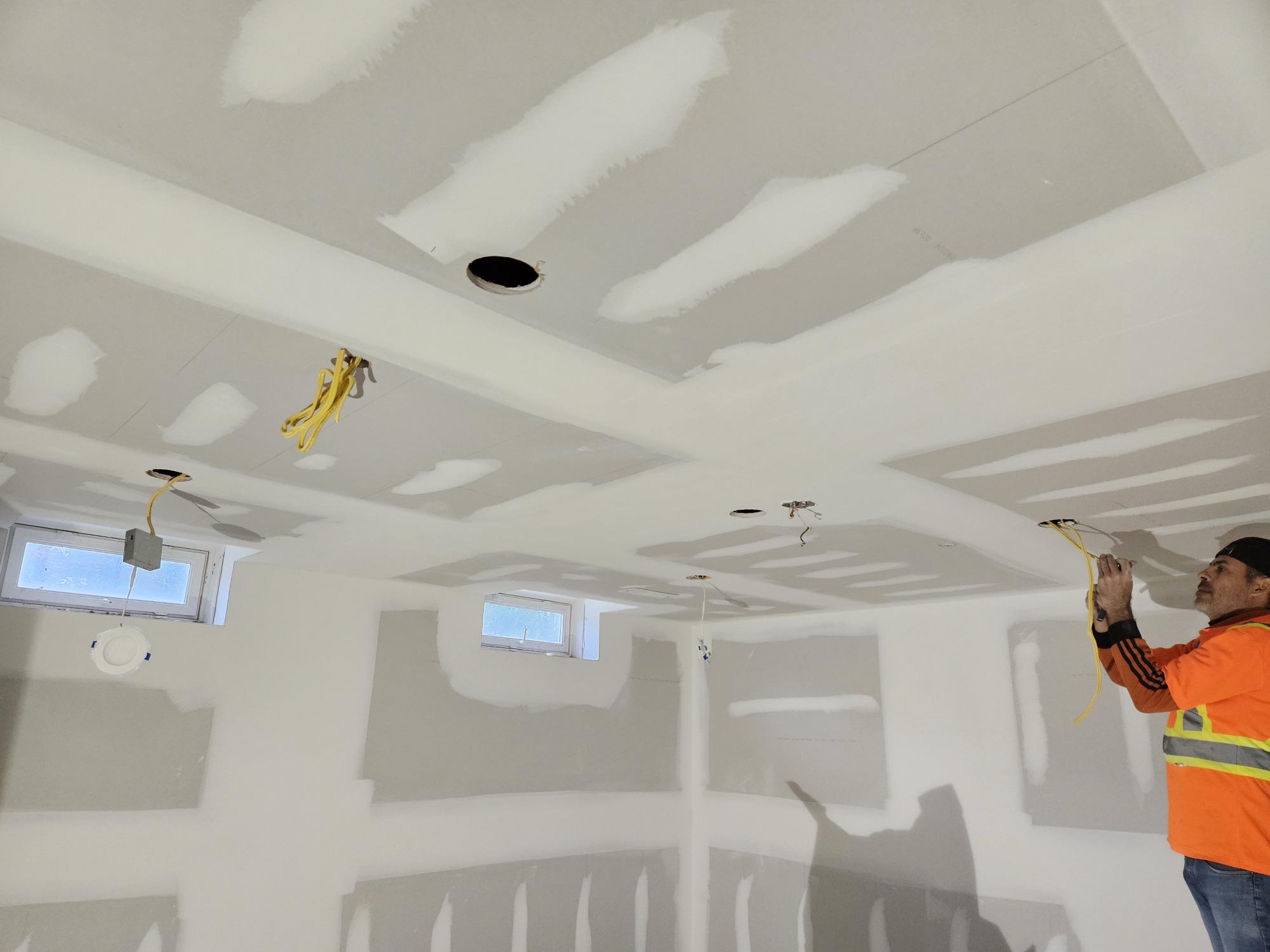 An electrician pulling wire through a freshly boarded and taped basement ceiling, pot-light holes cut and waiting for fixtures